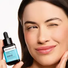 Uśmiechnięta kobieta trzyma serum z madekasozydem, które obiecuje naprawę niedoskonałości.