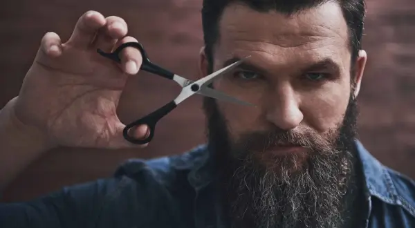 Mężczyzna z bujną brodą i nożyczkami przy oku, gotowy do modelowania brody.