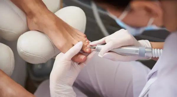 Profesjonalny pedicure frezarkowy w trakcie. Dłoń w białej rękawiczce używa frezarki do pielęgnacji paznokci stóp.