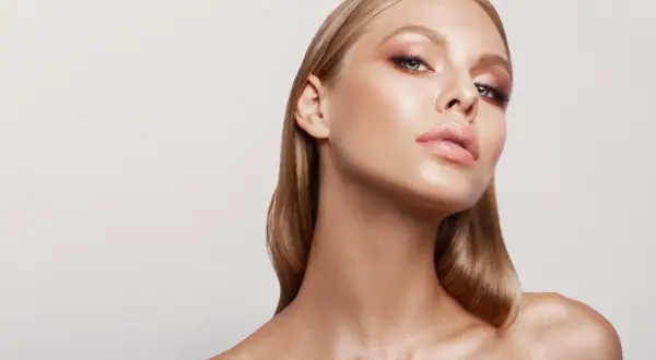 Blondynka z subtelnym makijażem, pokazująca, jak nakładać rozświetlacz na kości policzkowe i łuk kupidyna dla efektu glow.