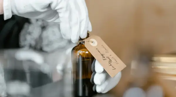 Białe rękawiczki trzymają buteleczkę z ciemnym płynem. Na etykiecie widnieje napis "Linalool".