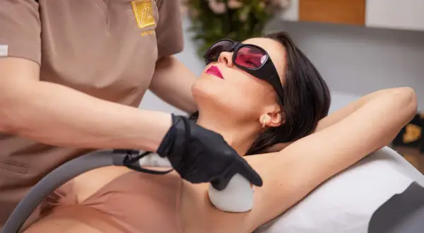 Kobieta w okularach ochronnych przechodzi zabieg depilacji laserowej pach. Czy depilacja laserowa boli? Zależy od indywidualnego progu bólu.