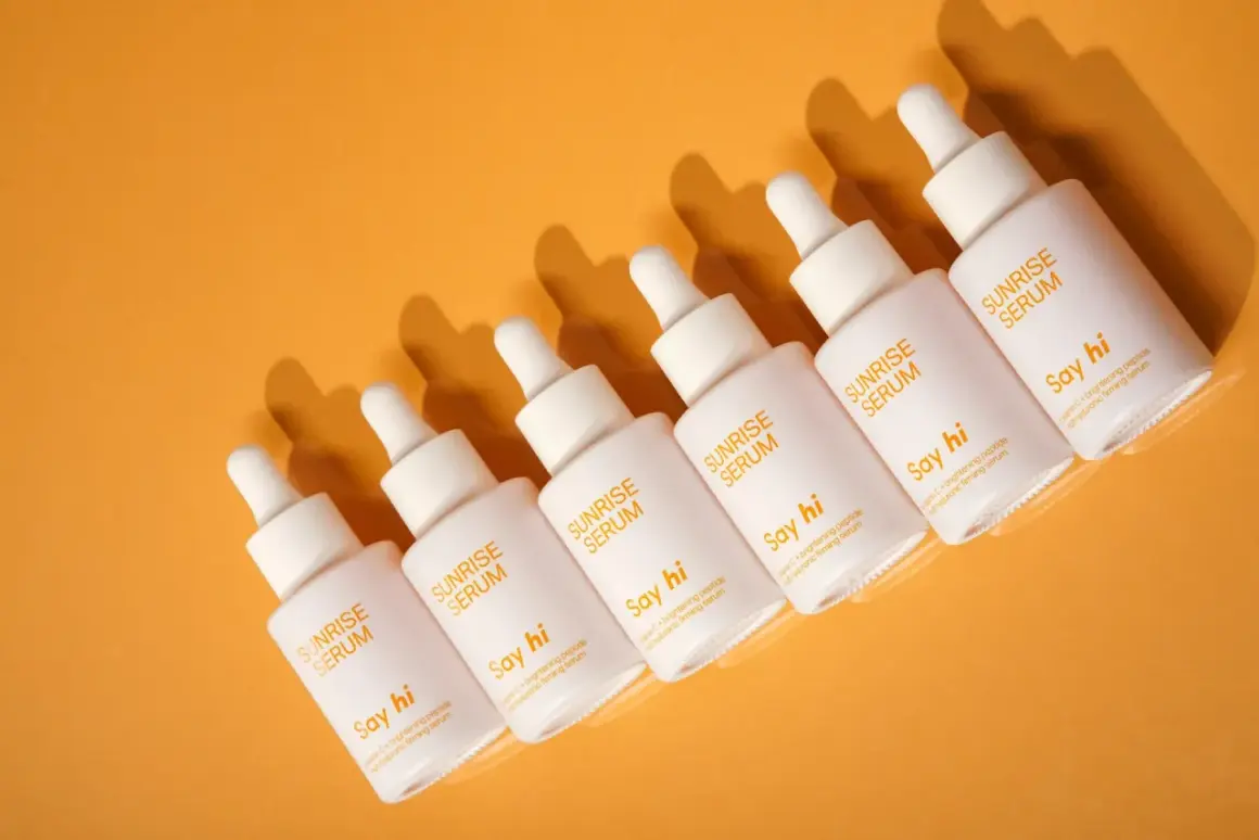Sześć białych buteleczek serum "Sunrise Serum" z pipetkami, z tekstem "Say hi" i "palmitynian askorbylu" na pomarańczowym tle.