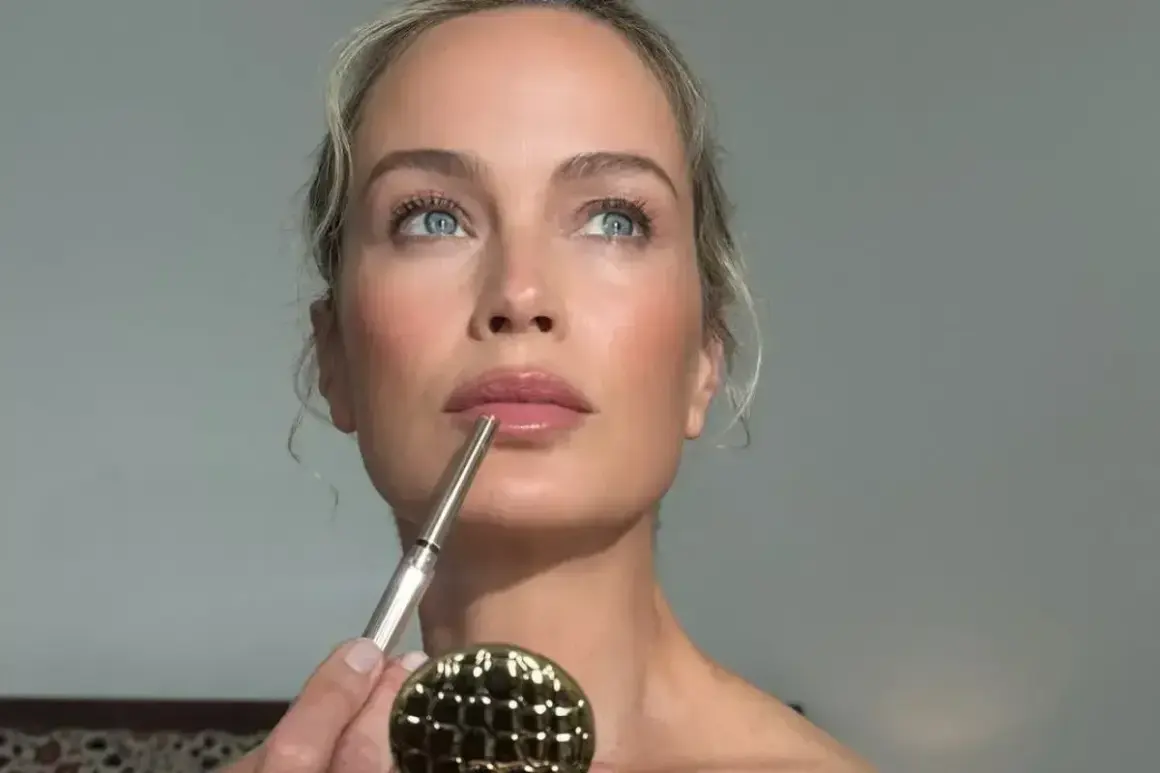 Kobieta z błękitnymi oczami i blond włosami wykonuje makijaż glamour, precyzyjnie obrysowując usta konturówką.