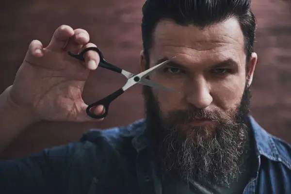 Mężczyzna z bujną brodą i nożyczkami przy oku, gotowy do modelowania brody.