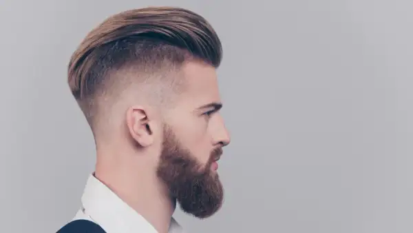 Mężczyzna z zarostem i fryzurą slick back, z wygolonymi bokami i dłuższymi włosami na górze.