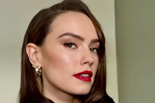 Elegancka kobieta z burzą brązowych włosów ułożonych w stylową side part fryzurę. Jej spojrzenie przyciągają zielone oczy i wyraziste, czerwone usta.