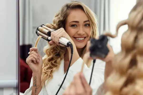 Uśmiechnięta blondynka w szlafroku tworzy fale prostownicą. Proste kroki, jak zrobić fale prostownicą, by uzyskać efektowną fryzurę.